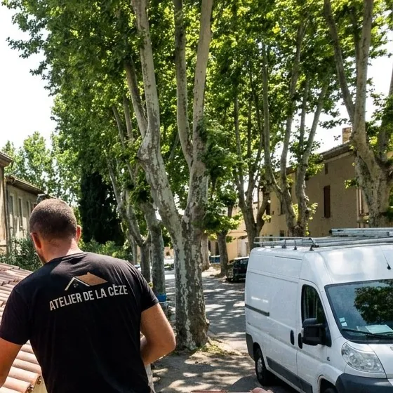 Couvreur de l'Atelier de la Cèze sur un toit à Bagnols-sur-Cèze, rue ombragée de platanes et camionnette garée
